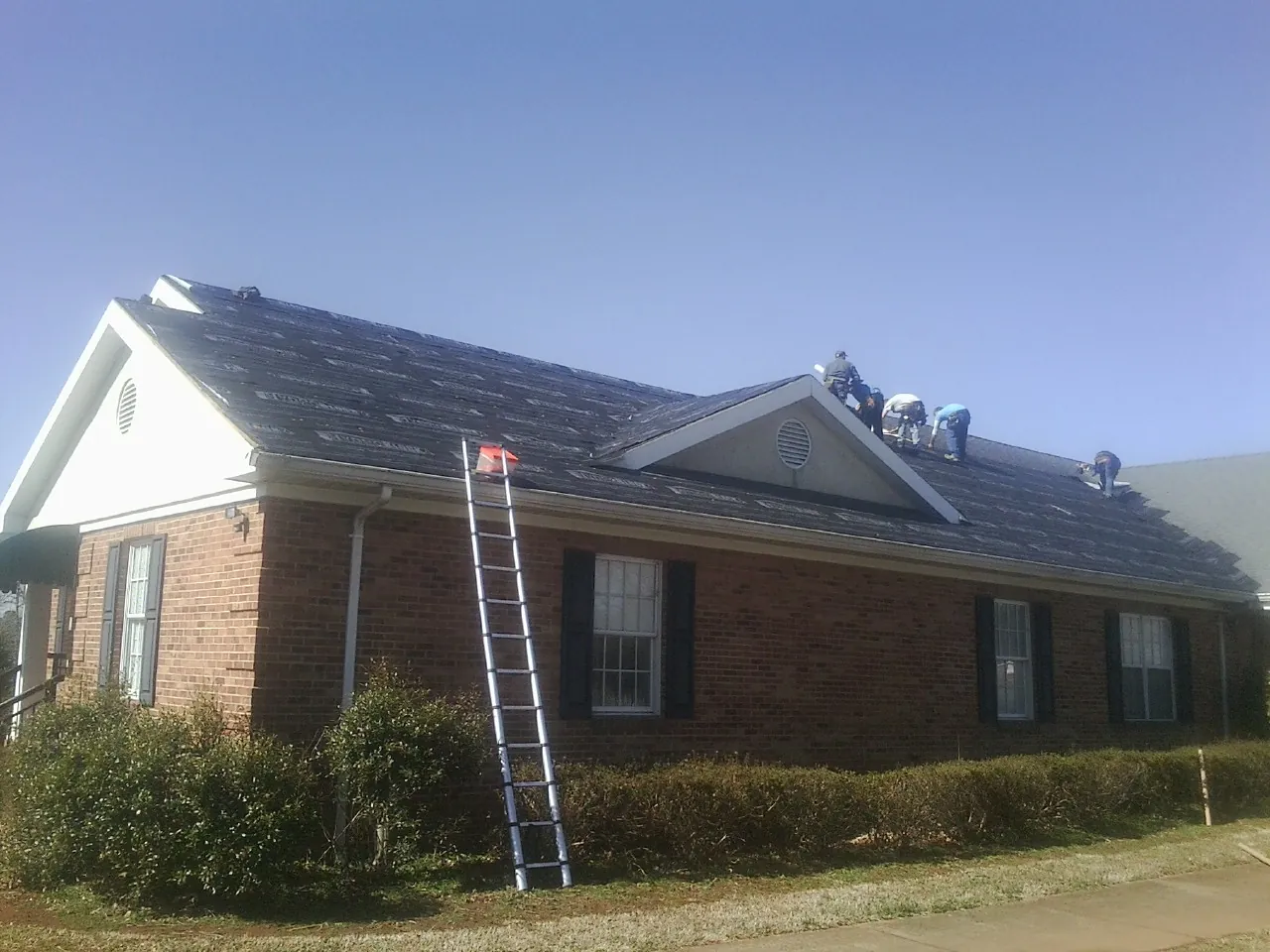 Crew tearing shingles off a brick ranch roof with a ladder set — Emergency Roof Repair work by our crew in East Moriches