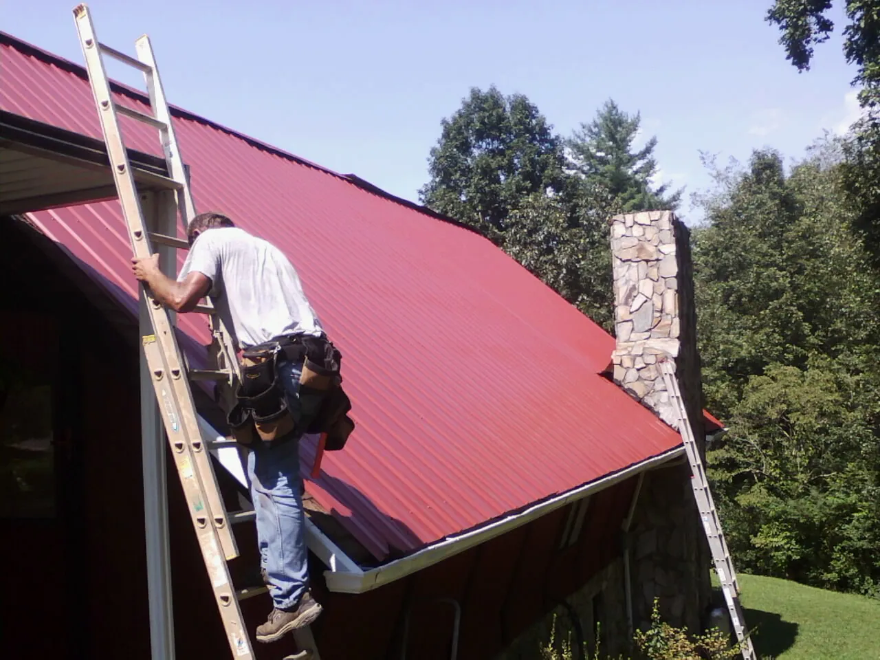 Roofer on a ladder beside a red metal roof and stone chimney — Soffit & Fascia Repair by Roofing Brothers in East Moriches